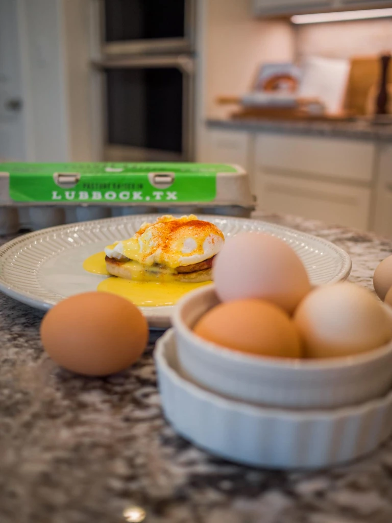 Photo of eggs benedict on a plate with fresh Alcove eggs in the foreground and a carton of Alcove eggs in the background.