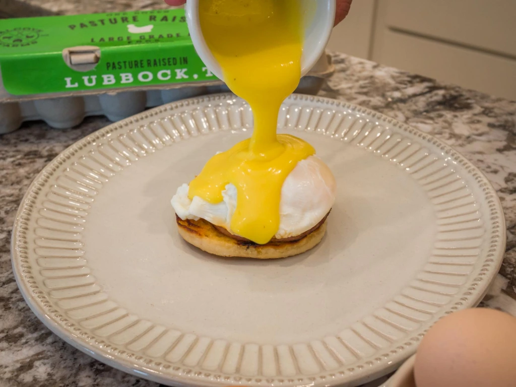 Photo of fresh hollandaise sauce being poured over a poached egg. A carton of Alcove Farms eggs can be seen in the background.