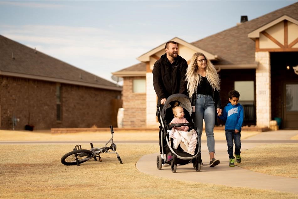 Photo of a family walking in front of their house. Dad pushes a baby girl in a stroller, and mom walks next to a son.