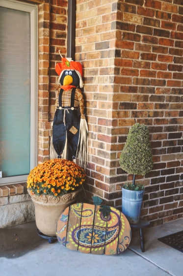 Photo of a Betenbough home front porch decorated with a scarecrow, pot of mums, painted pumpkin, and topiary.