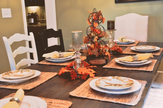 Photo of a dining table set with straw place mats and fall-themed dishes. The centerpiece is pumpkins, leaves, and candles.