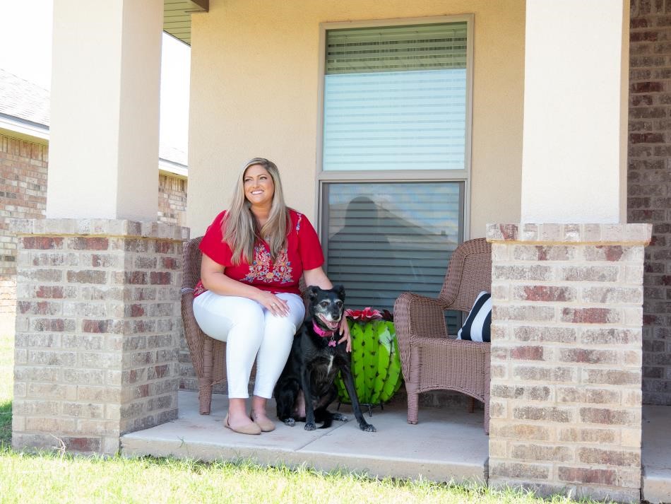 Photo of a woman on the front porch of her new Betenbough house with her pet, a black dog with a graying face.