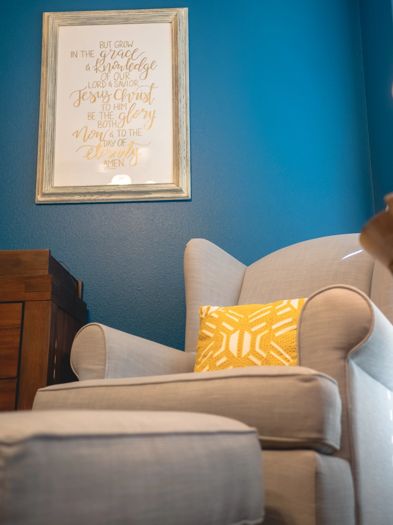 Photo of nursery decor that shows blue walls, a window, a gray chair and ottoman, a side table with a lamp and clock, a changing station, and a laundry hamper.